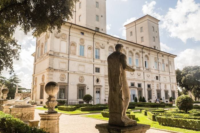 The facade of the Galleria Borghese seen across its front lawn, with stone statues lining the approach