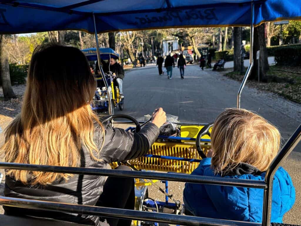 Two kids pedaling a risciò pedal cart along a paved path in Villa Borghese